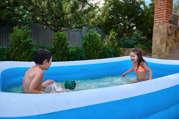 Water fun day in a backyard. Happy children playing ball, splashing each other with water in inflatable swimming pool