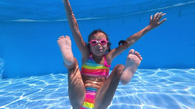 Little girl in color bikini and pink glasses dive under water in pool. Child holds breath and sits in lotus position. Sport good for healthy. Having fun in the pool. Vacation and hobby.