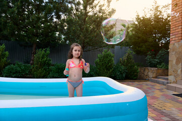 Charming little girl catches soap bubbles that she blows in inflatable water pool at home garden on a sunny summer day