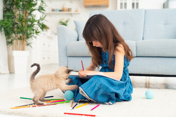 Child girl painting with kitten and lying on the floor. Little female person drawing with colorful pencils and playful kitty pet next to her at home playing with a ball