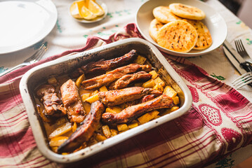 grilled pork ribs on table inside oven aluminum container with venezuelan arepas and potatoes, table cloth, and warm natural light