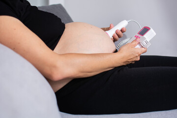Pregnant woman with a fetal doppler listening to the baby's heart sitting on the sofa at home	