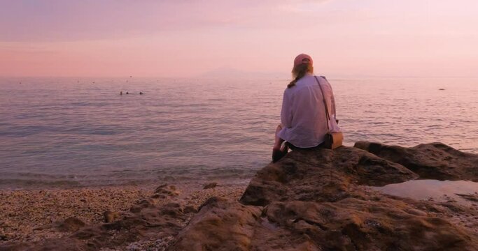 Visually attractive scene of lonely young woman sitting on rocks beach enjoy stunning sunset light over sea, back view. Summer holidays.