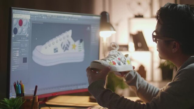 Close Up Of Asian Male Footwear Designer Looking At The Colourful Pattern Sneaker In Hands And Comparing It To The Pictures On A Desktop At Home. Shoe Production Procedure Concept 
