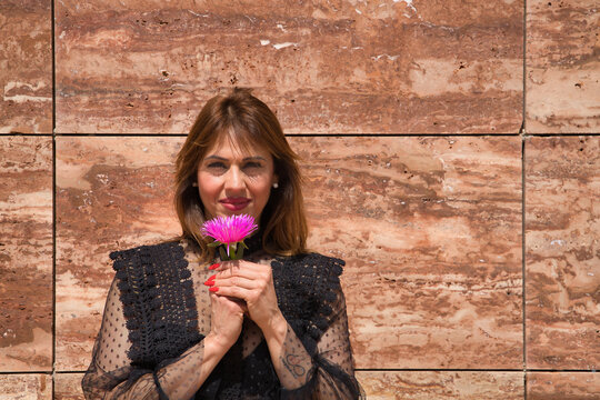 Portrait Of Attractive Mature Woman In Transparent Black Shirt, Smelling A Beautiful Flower On A Brown Marble Background. Concept Maturity, Beauty, Makeup, Wrinkles, Flowers.