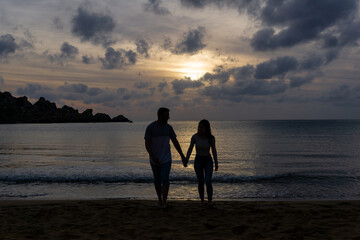 Silhouette of two lovers holding hands and walking on the beach with the sunset in the background.