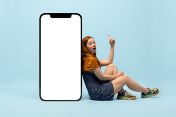 Young happy girl next to huge 3d model of smartphone with empty white screen isolated on blue...