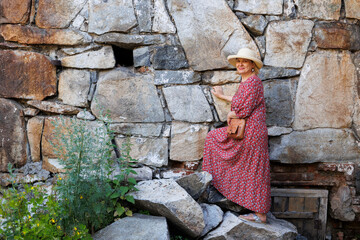 A stylish aged woman in a folklore dress and a straw hat on the background of a stone wall. Summer fashion for older women. - 516951872