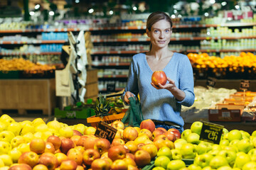 woman picks chooses fruits vegetables on the counter in supermarket. Female housewife shopping in market standing near vegetable department store with a basket in hands. Examines apple