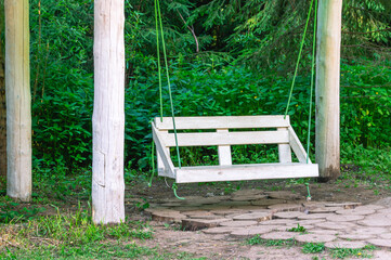 White wooden swing in the forest next to the trees. Entertainment for children and adults in the park. A place to relax and unwind.
