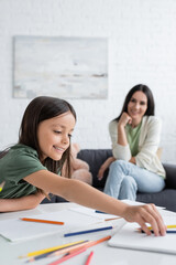 happy girl reaching colorful pencils near papers on table and blurred babysitter on background.