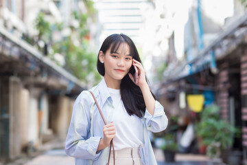 Confident young Asian woman who wears a blue white shirt and bag smiles happily while she holds talks to customer on the phone with smartphone as she commute to work through the old town.