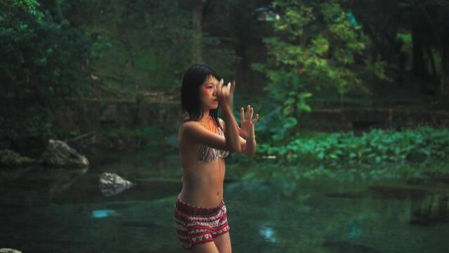 Elegant Asian girl with painted body performing an extatic dance in a lush tropical forest near calm clean pond. 