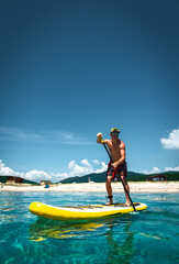 Tanned man stands on a yellow supboard and paddles. Active sport on the beach in the turquoise sea...