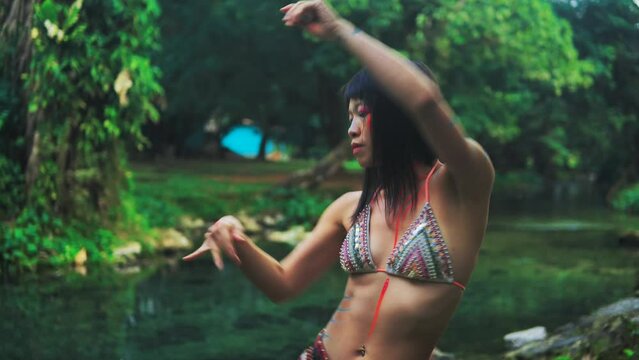 Elegant Asian girl with painted body performing an extatic dance in a lush tropical forest near calm clean pond. 