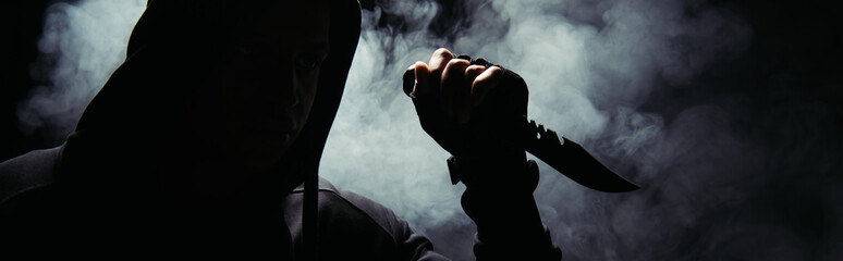 Silhouette of man in hoodie holding knife on black background with smoke, banner