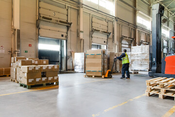 Interior of a modern warehouse storage of retail shop with pallet truck near shelves and loading ramps. Storehouse worker in uniform working on forklift.