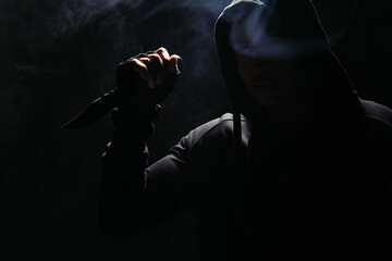Silhouette of african american bandit in hood holding knife on black background with smoke