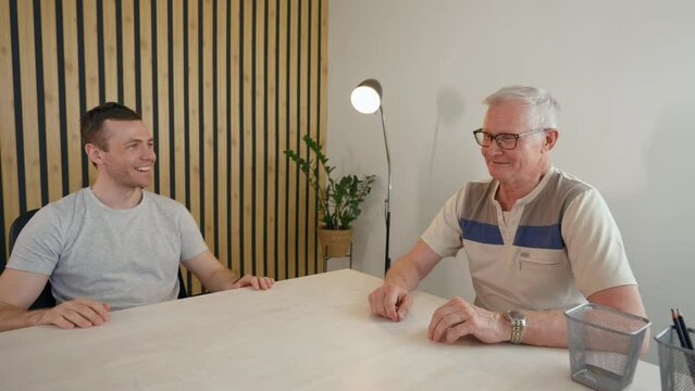Young businessman talks to elderly client discussing new joint project. Man sits at desk in office turning to camera to summarize from third party to POV