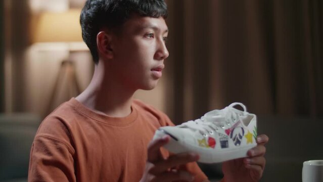 Close Up Of Asian Boy Footwear Designer Holding The Colourful Pattern Sneaker And Comparing It To The Pictures On A Desktop Computer At Home
