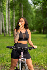 Fototapeta na wymiar A beautiful girl with tattoos in a sports uniform rides a bike and eats ice cream
