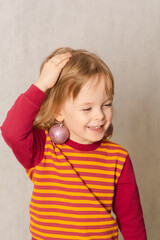 Smiling child put Christmas balls on his ears like earrings. Girl playful and cheerful at christmas holiday. Real people, simple life. Vertical