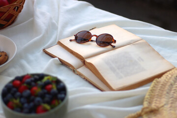 Picninc blanket with straw bag, bowl of strawberries and blueberries, bowl of chocolate chip cookies, books, sunglasses and basket of apples in the garden. Selective focus.