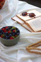 Picninc blanket with straw bag, bowl of strawberries and blueberries, bowl of chocolate chip cookies, books, sunglasses and basket of apples in the garden. Selective focus.