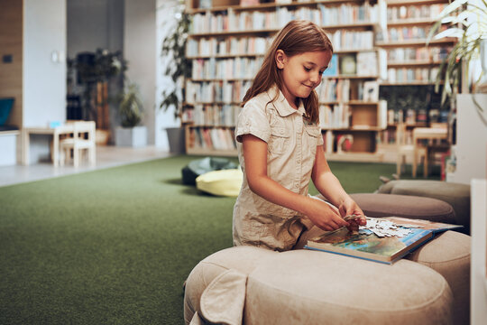 Schoolgirl doing puzzle and reading book in school library. Primary school pupil is involved in book with jigsaw. Benefits of everyday reading. Child curiosity