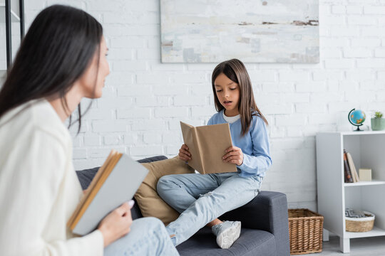 girl sitting on couch and reading book near blurred babysitter.