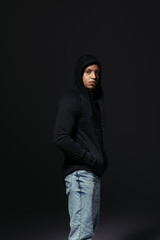 African american hooligan holding hand in pocket of hoodie isolated on black