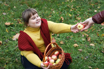 Obese girl with red scarf sits on grass and holds basket with apples in autumn