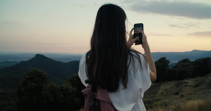 Beautiful young adult woman making photo or video using smartphone stand on top of the mountain on summer hike journey. Female influencer or blogger making digital content using mobile phone. 