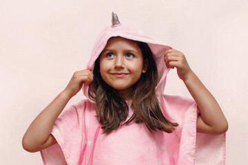 a little girl with a smile in a pink poncho towel looks up, a child in a unicorn costume on a white...