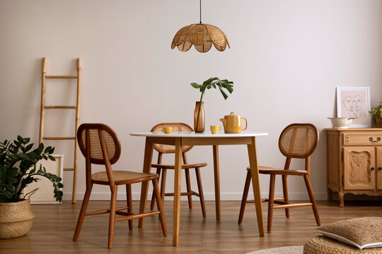 The stylish dining room with round table, rattan chair, wooden commode, pock up poster and kitchen accessories. Beige wall with mock up poster. Home decor. Template.