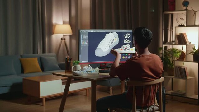 Asian Boyfootwear Designer Looking At The Colourful Pattern Sneaker In Hands And Comparing It To The Pictures On A Desktop At Home. Shoe Production Procedure Concept 
