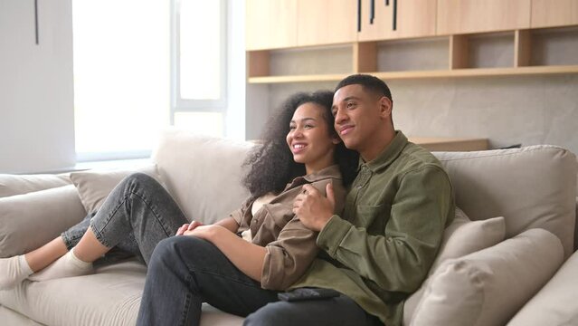 Side view of the multiracial couple chilling on the couch, watching movie in the studio apartment. Guy switching channel with TV remote