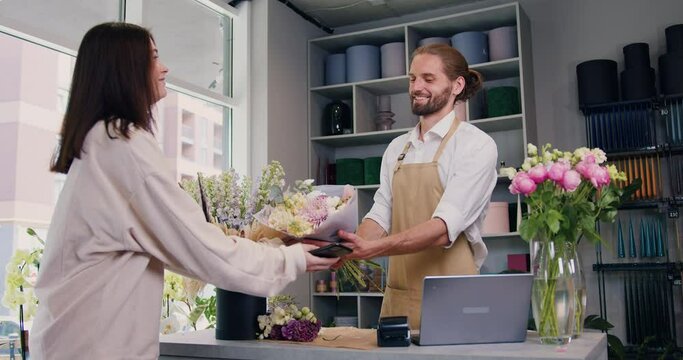 Woman buying flower composition and paying by credit card using terminal. Man florist sells a bouquet of flowers to a happy woman, a female buyer pays. Small business.