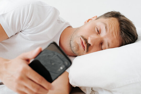 Sleepy tired young man woke up in morning from an alarm clock on mobile phone. Lazy guy holding smartphone while lying in bed in bedroom and looking at screen
