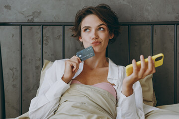 Young minded woman wear white shirt pajama she lying in bed use mobile cell phone credit bank card...