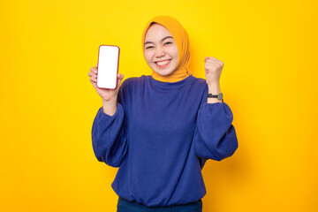 Excited young Asian Muslim woman dressed in casual sweater showing blank screen mobile phone and making winner gesture isolated over yellow background