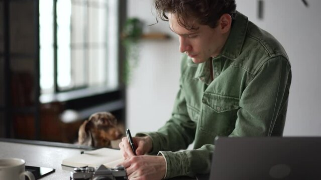 4k Young man is writing in notebook and sitting at table with dog at home spbd. Close view of handsome caucasian guy takes notes and looks with smile, uses laptop and sits at desk with cute pet in
