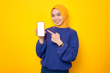 Smiling young Asian Muslim woman dressed in casual sweater showing blank screen mobile phone, recommending mobile app isolated over yellow background