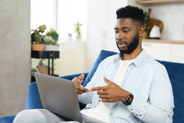 Serious concentrated male student involved virtual meeting, webinar. Black man making video call on the laptop, sitting on the sofa at home, talking and gesturing, explaining something