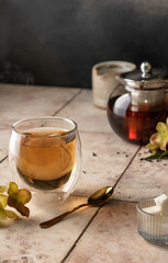 Tea in thermal glass with steam on beige tile background in composition with flowers, sugar and teapot. Text space