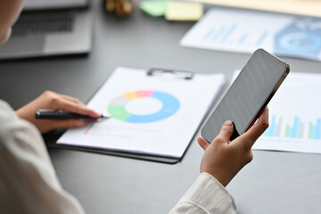 Selective focus on Businesswoman's hand holding smartphone, checking, examining and analyzing financial diagrams show on paperwork.