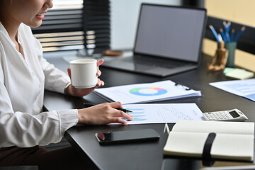 Cropped shot of Asian Businesswoman sitting in the office and holding a cup of coffee while checking, examining and analyzing financial report.