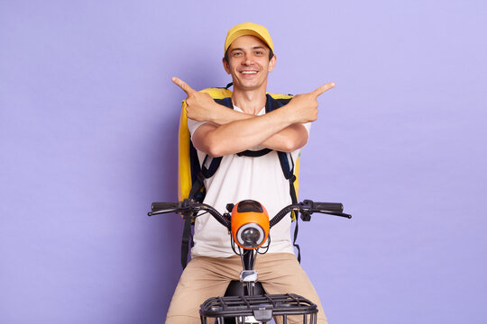 Image of young courier man of takeaway food on electric scooter posing isolated over purple background, showing x sign, pointing to both sign, presenting copy space for advertisement.