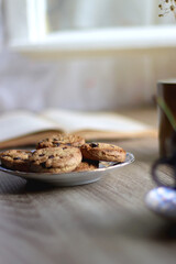 Plate of chocolate chip cookies, cup filled with strawberries, blueberries and cherries, open book and vase with gypsophila flowers on the table. Selective focus.