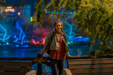 A woman stands under a green tree near a pond with bright glowing colored jets of a fountain in a nighttime amusement park. Blurred images